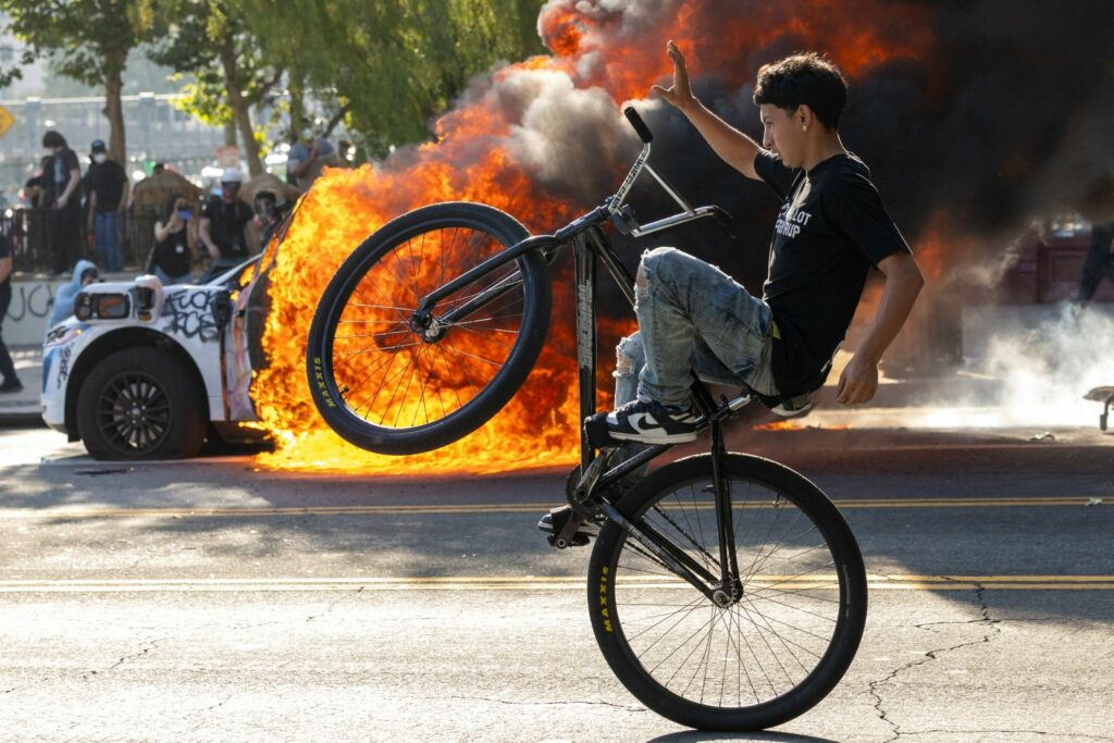 Young person doing a wheelie on a bicycle. Background has a car in flames.